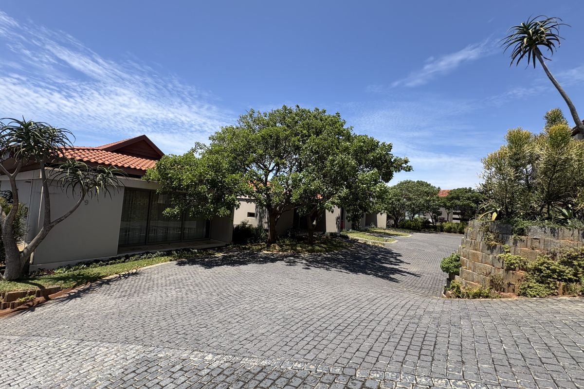 A well-maintained front exterior view featuring a cobblestone driveway, mature trees, and a residence with terracotta roof tiles and large sliding glass doors.