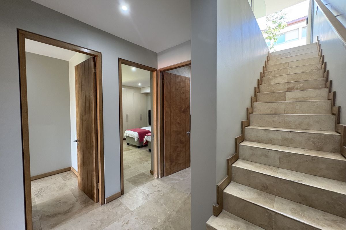 A bright, modern hallway featuring stone tile flooring, a timber staircase, and solid timber doors leading to adjacent rooms.