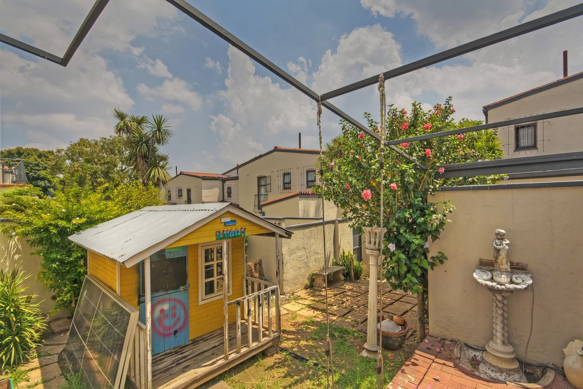 A rear garden area featuring a small wooden play structure, paved pathways, and perimeter boundary walls within a townhouse complex setting.