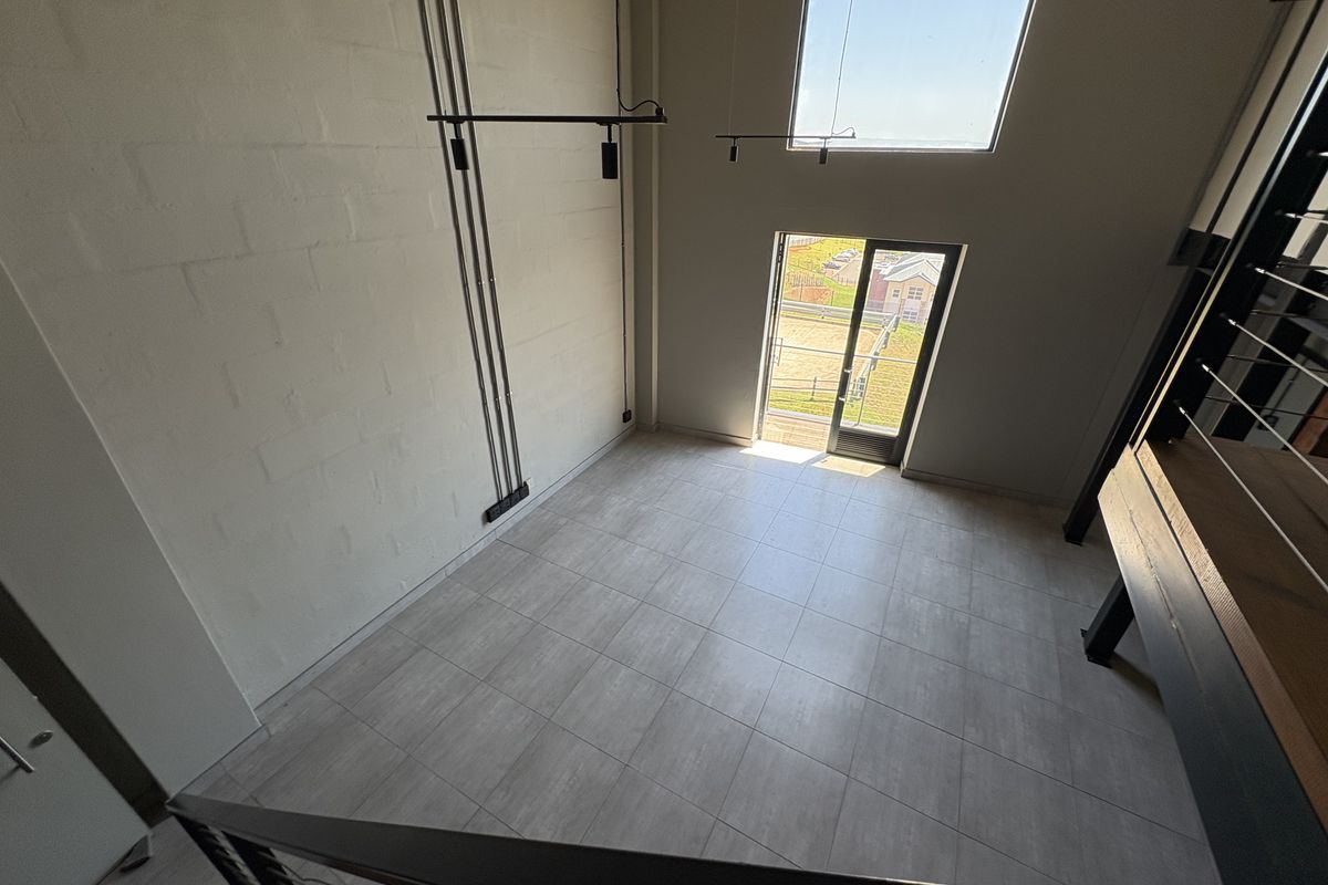 A bright, double-volume living area featuring modern tiled flooring, exposed industrial-style electrical conduits, and large windows providing natural light and outdoor access.