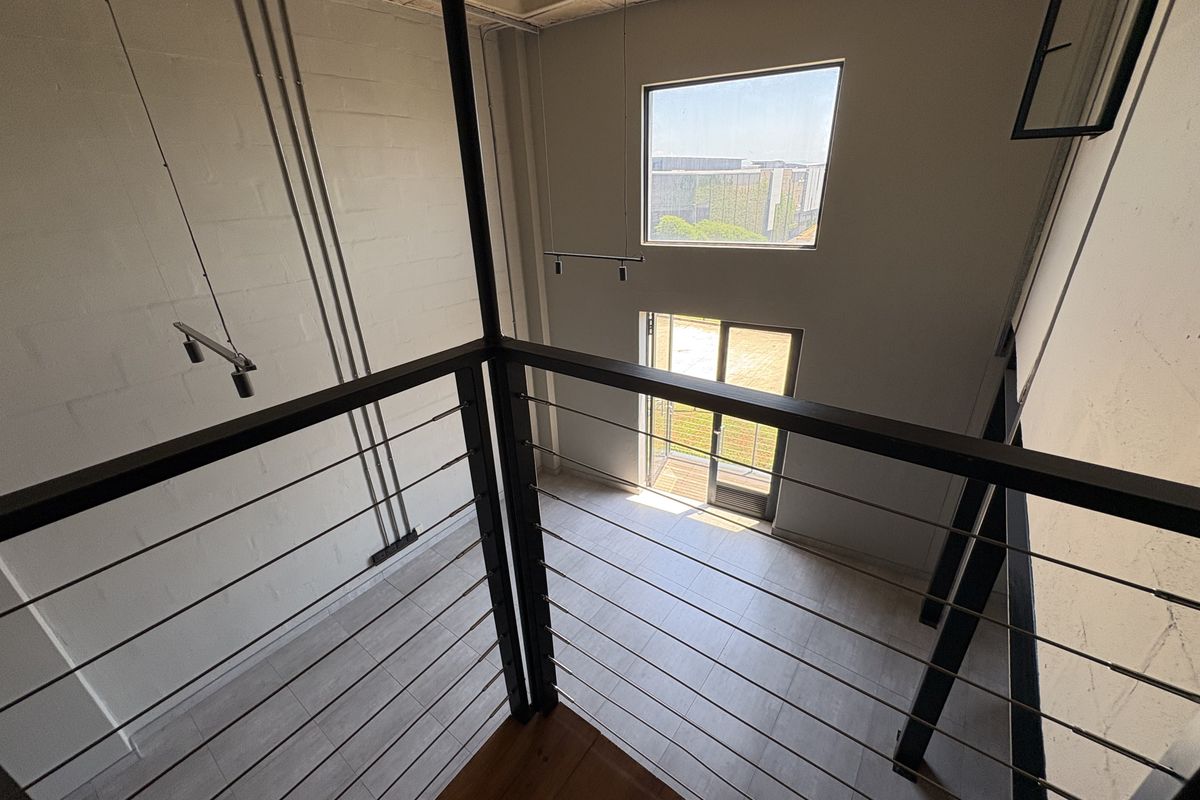 A double-volume living area featuring industrial-style exposed concrete ceilings, metal balustrades, and large windows providing natural light and access to an outdoor space.