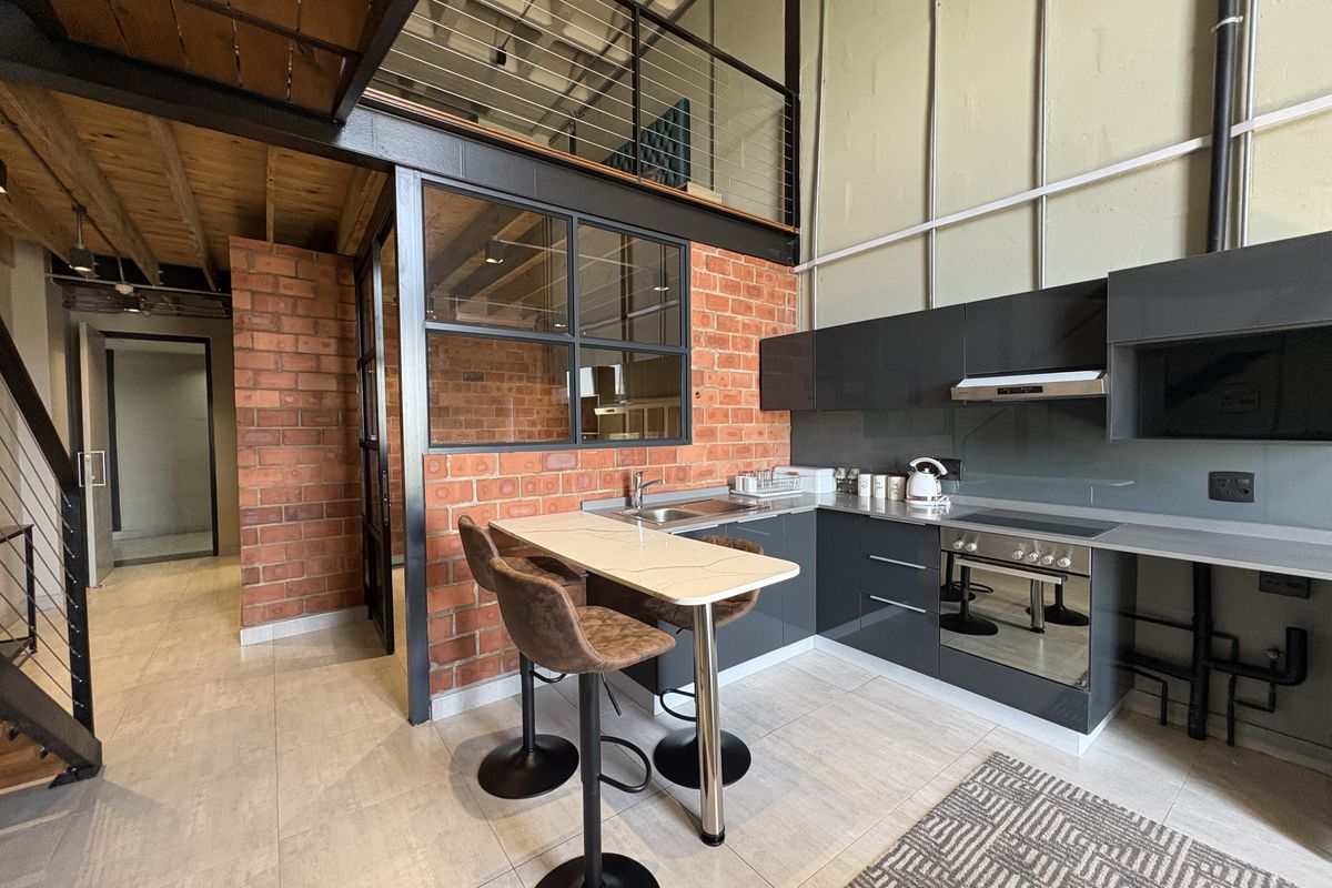 Modern industrial-style kitchen featuring exposed brick walls, sleek dark cabinetry, integrated oven and hob, and a breakfast bar with a mezzanine level above.