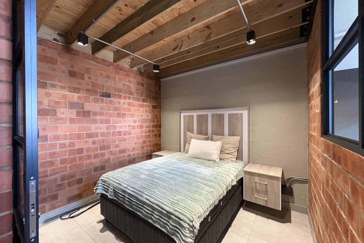 Industrial-style bedroom featuring exposed brick walls, timber beam ceiling, tiled flooring, and large steel-framed windows.