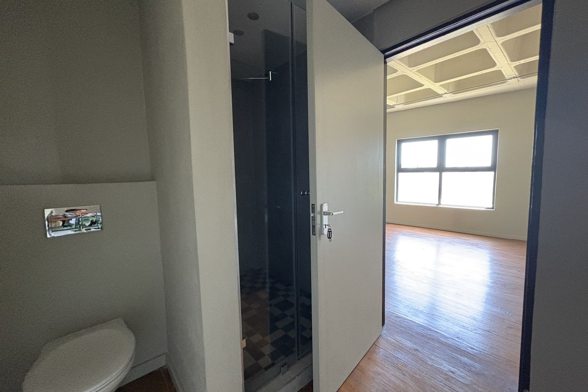 A modern bathroom featuring a wall-mounted toilet and a glass-enclosed shower, with a view into an adjacent open-plan living area with wooden flooring.