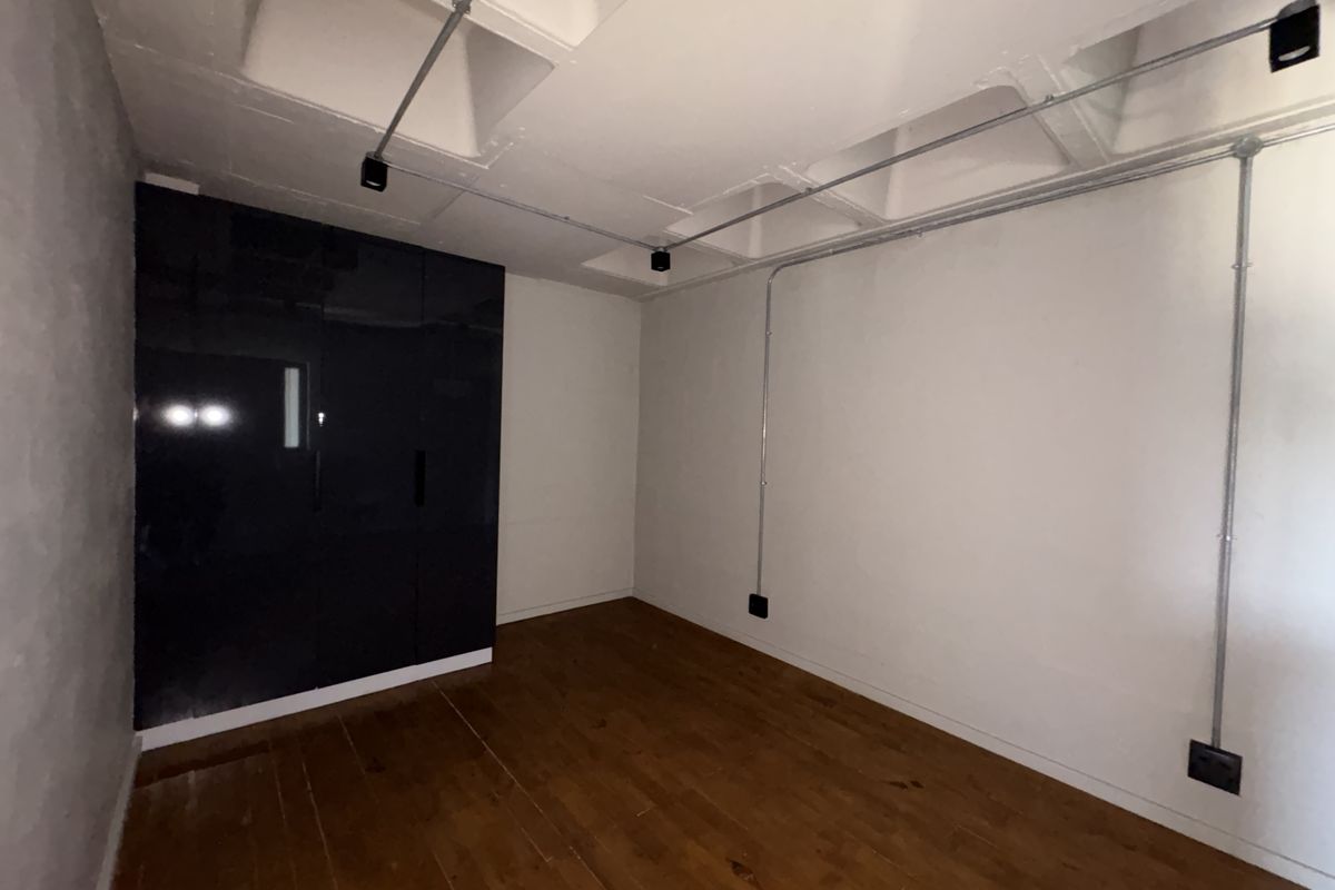 A clean, empty bedroom featuring dark built-in cabinetry, exposed conduit lighting, and warm-toned wooden flooring.
