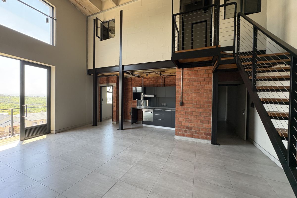 Spacious industrial-style open-plan living area featuring exposed brickwork, high coffered ceilings, a modern kitchen, and a mezzanine level with a staircase.