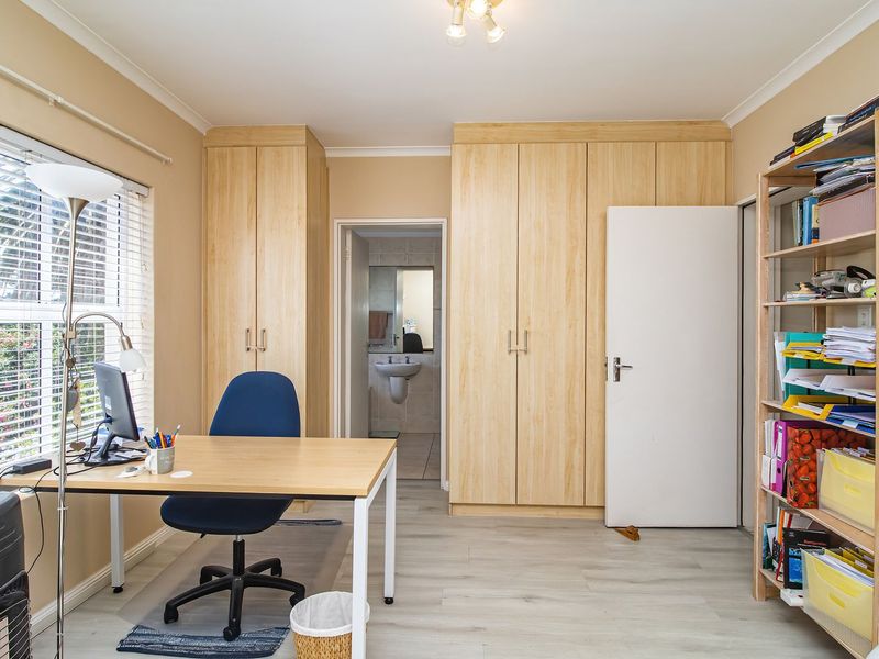 A functional bedroom featuring light wood-finish built-in cupboards, laminate flooring, and large windows with venetian blinds, offering direct access to an en-suite bathroom.
