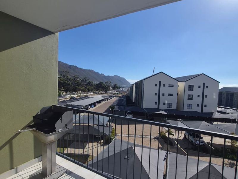 A covered balcony featuring tiled flooring, a metal balustrade, and a built-in braai, offering views over the apartment complex parking area.