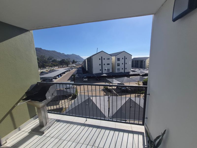 A covered balcony featuring tiled flooring, a metal balustrade, and a built-in braai, offering views over the apartment complex parking area.