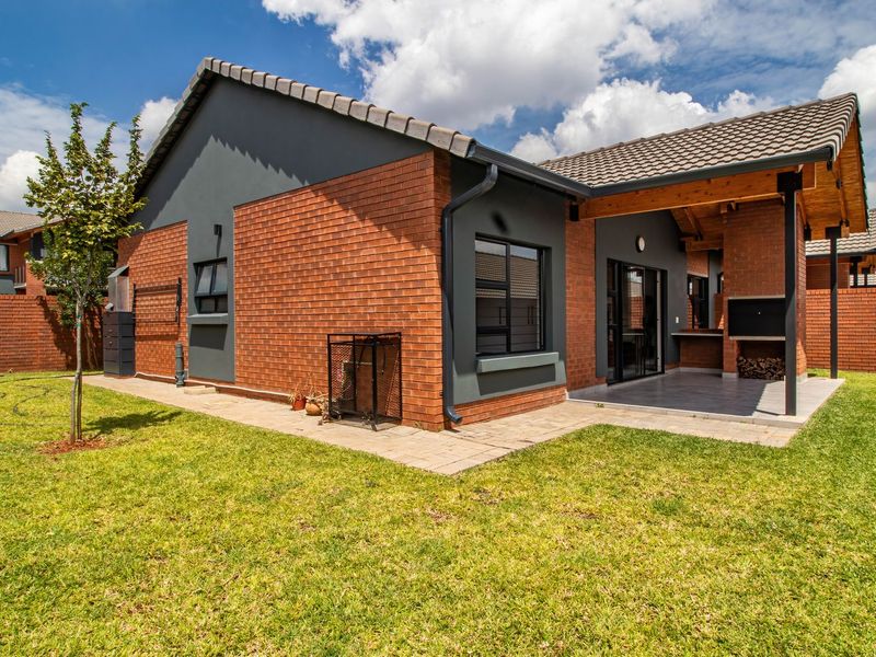 A well-maintained townhouse rear exterior featuring a covered patio with a built-in braai, brick facade, tiled roof, and a private enclosed garden.