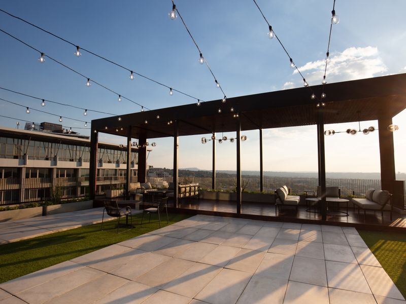 A spacious rooftop terrace featuring a covered pergola, integrated string lighting, paved flooring, and artificial turf, offering expansive urban views.