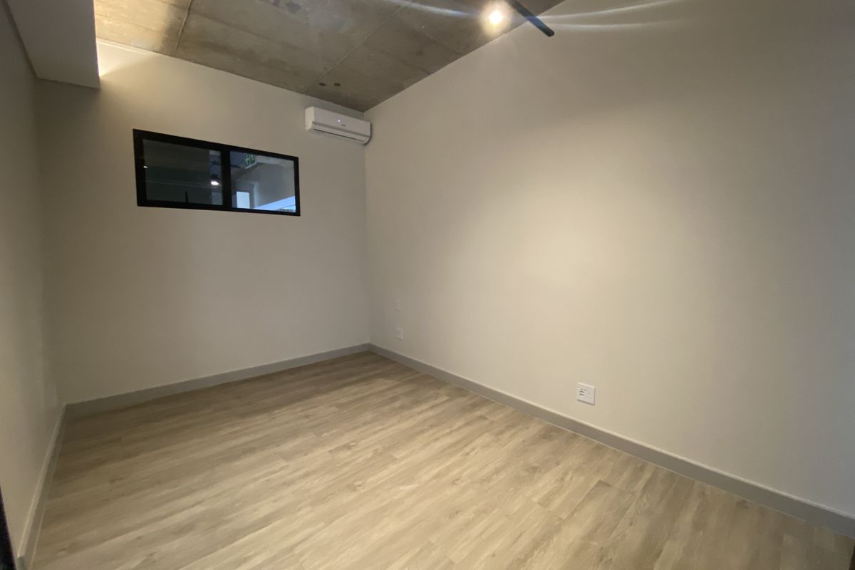 A modern, empty bedroom featuring light wood-look flooring, an exposed concrete ceiling, and a wall-mounted air conditioning unit.