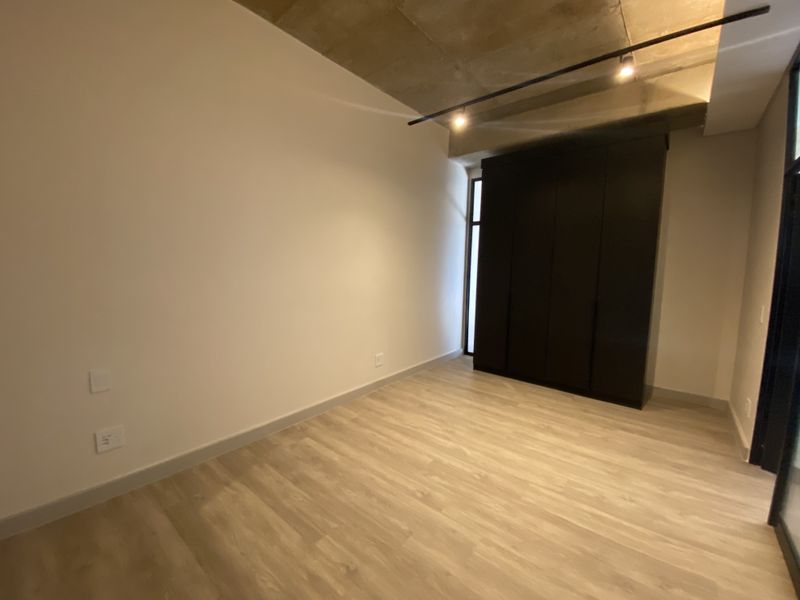A modern bedroom featuring light wood-look flooring, a dark built-in wardrobe, and an exposed concrete ceiling with track lighting.