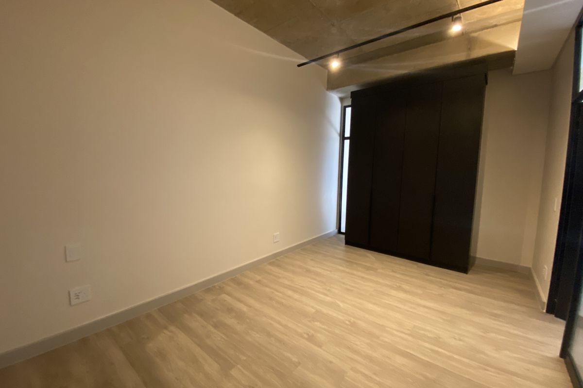 A modern bedroom featuring light wood-look flooring, a dark built-in wardrobe, and an exposed concrete ceiling with track lighting.