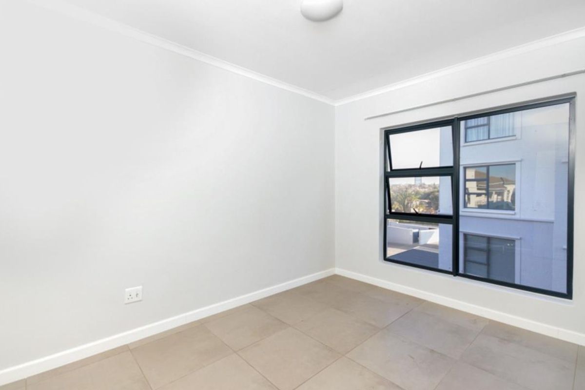 A bright, empty bedroom featuring neutral tiled flooring, clean white walls, and a large window with dark framing providing natural light.