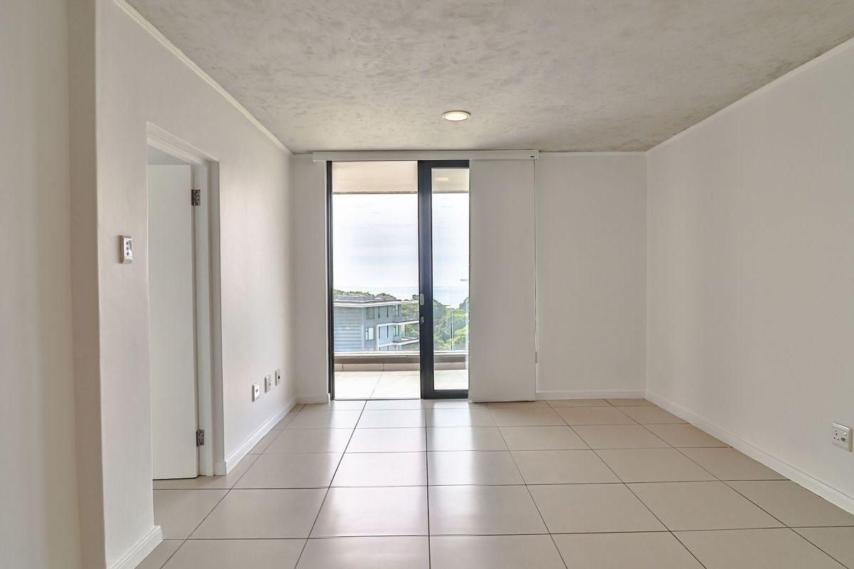 A bright, empty living area featuring neutral floor tiles, recessed ceiling lighting, and a sliding glass door leading to an outdoor balcony.