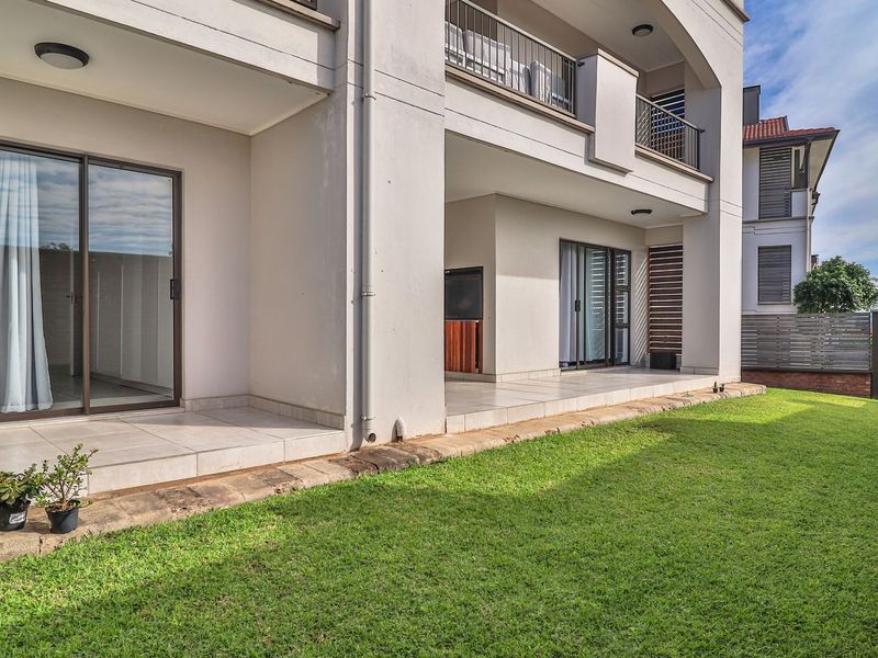 A well-maintained rear exterior view of an apartment featuring a spacious tiled patio, sliding glass doors, and a private landscaped garden area.