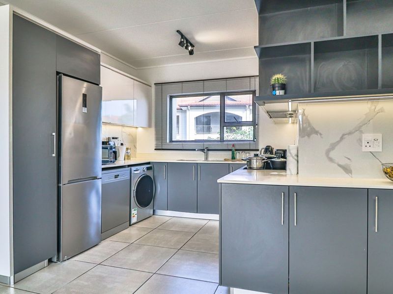 Modern kitchen featuring sleek grey cabinetry, stone countertops, marble-look splashback, integrated sink, and contemporary track lighting with tiled flooring.