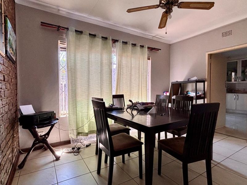 Dining room featuring tiled flooring, an exposed brick feature wall, and a ceiling fan, with an adjacent view into the kitchen area.