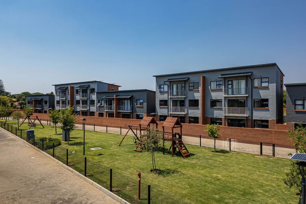 Exterior view of a modern apartment complex featuring landscaped communal gardens, a children's play area, and paved driveway access.
