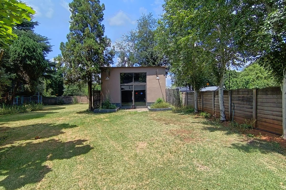 A spacious rear garden featuring a detached outbuilding with large glass doors, mature trees, and perimeter fencing.