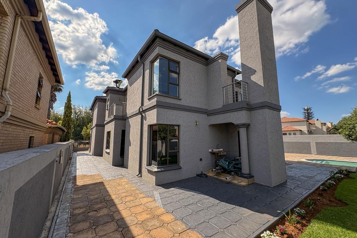 A view of the rear exterior of the double-story house, featuring a paved patio area, a tall chimney stack, and a balcony.