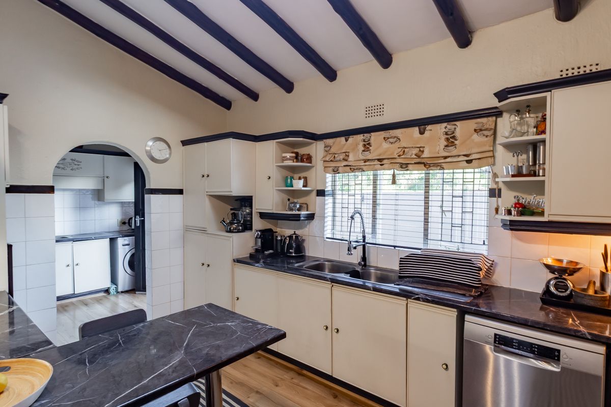 A functional kitchen featuring dark marble-look countertops, cream cabinetry, exposed ceiling beams, and an adjoining scullery area with laundry facilities.