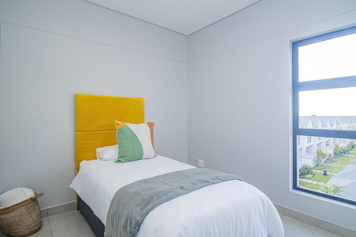 A bright and modern bedroom featuring neutral walls, tiled flooring, and a large window providing natural light and views of the surrounding complex.