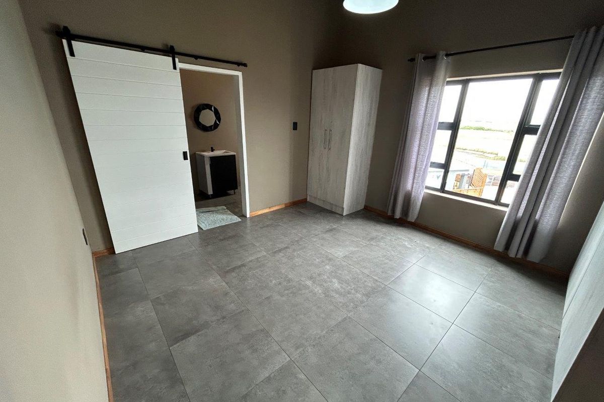 A bedroom featuring large format grey floor tiles, a sliding barn door leading to an en-suite bathroom, and a window with curtain tracks.