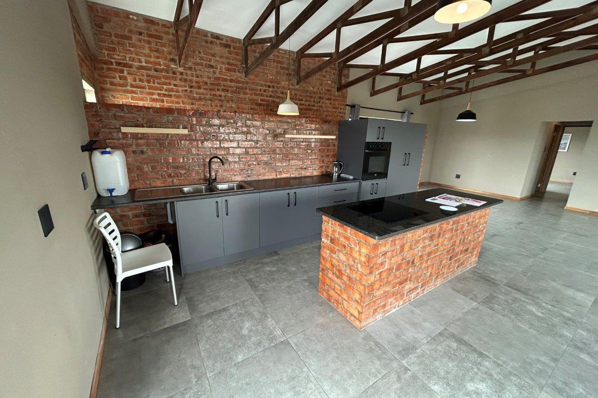 Modern industrial-style kitchen featuring exposed brick walls, dark grey cabinetry, a central island with a cooktop, and high vaulted timber truss ceilings.