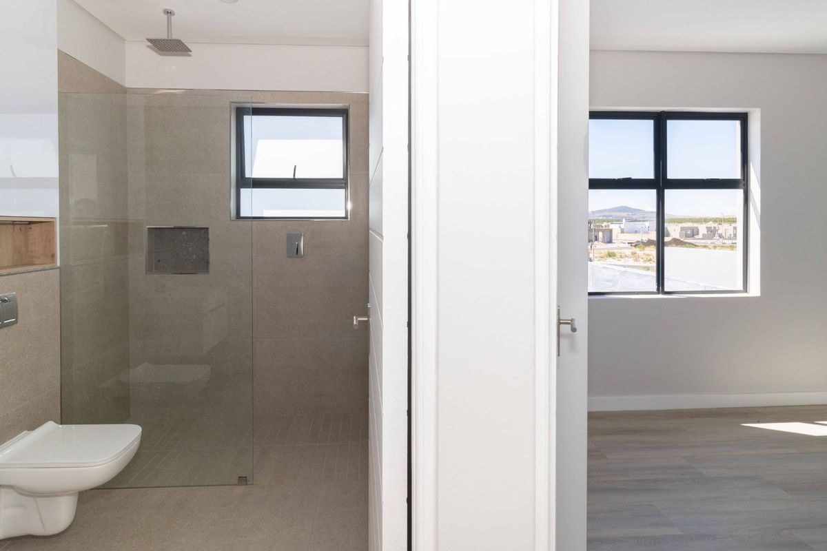 A modern bathroom featuring a frameless glass shower and wall-mounted toilet, adjacent to a bright, empty bedroom with neutral flooring.