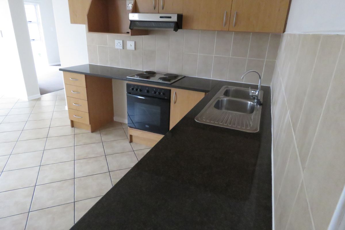 A functional kitchen featuring light wood cabinetry, dark countertops, a built-in oven with hob, and tiled flooring, presented in a clean, empty state.