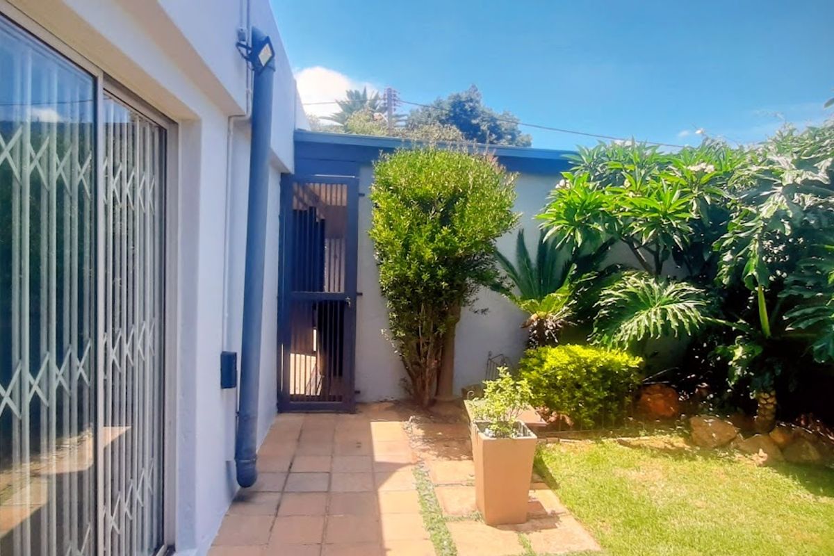 A private rear garden area featuring a paved walkway, established greenery, and secure access via a sliding door with security gate.