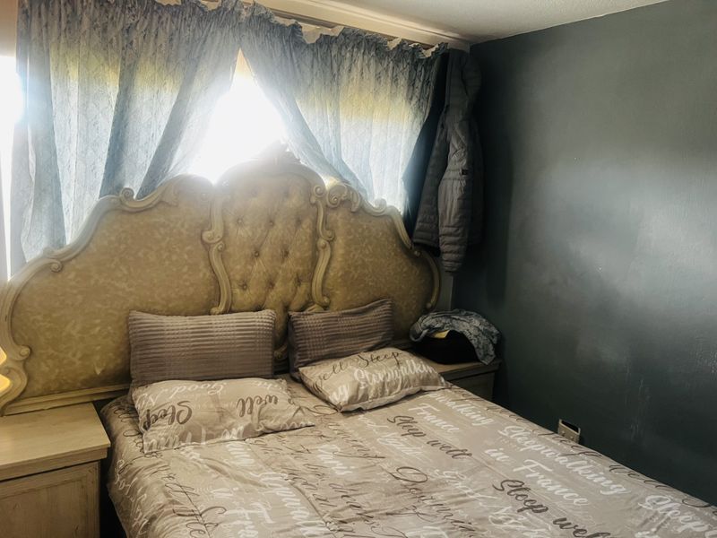 A bedroom featuring a large ornate headboard, neutral wall tones, and natural light filtering through window curtains.