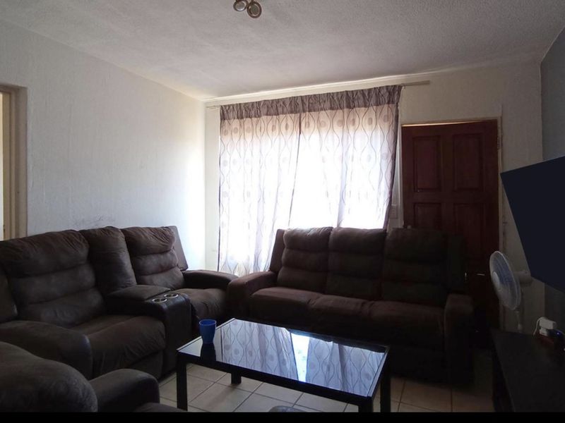 A living room featuring tiled flooring, a wooden door, and a large window with curtains, providing a functional and comfortable living space.