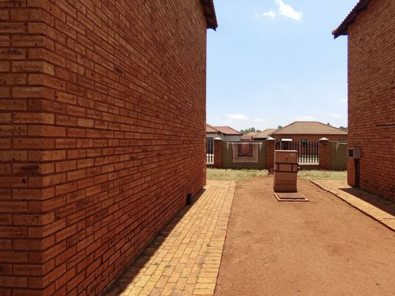 Exterior view showing the side passage between two brick apartment buildings with paved walkways and a fenced boundary wall in the background.