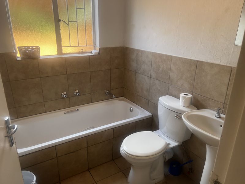 A functional bathroom featuring a built-in bathtub, pedestal basin, and toilet with neutral wall tiling and a security-grilled window.