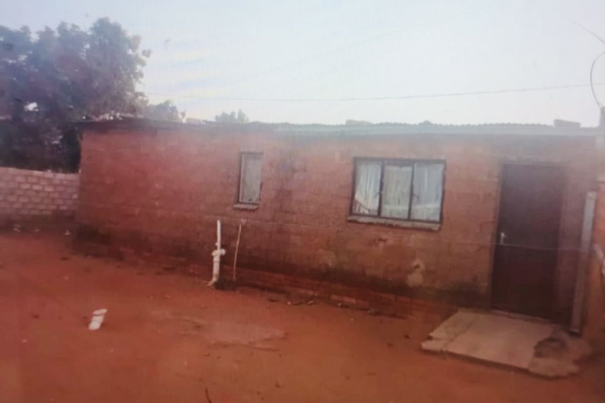 Exterior view of a single-story brick structure with a basic window and door, set on an unpaved plot.