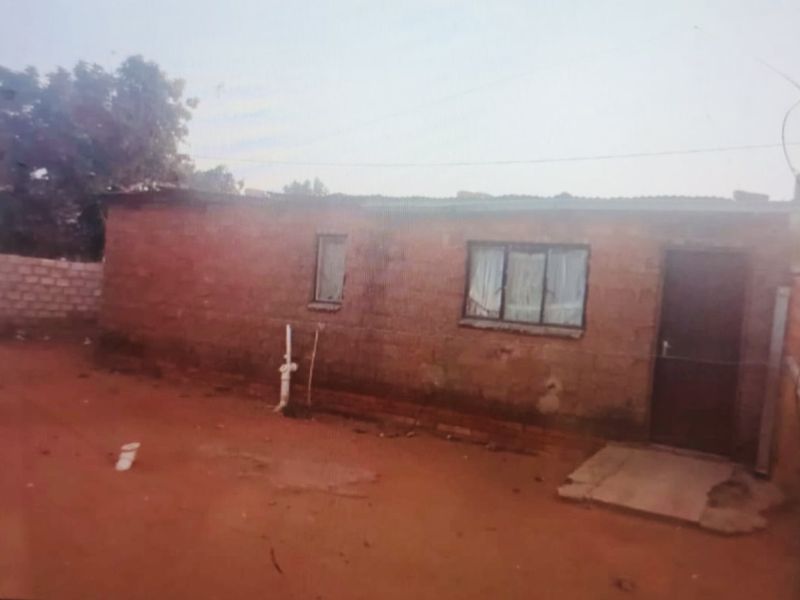 Exterior view of a single-story brick structure with a basic window and door, set on an unpaved plot.