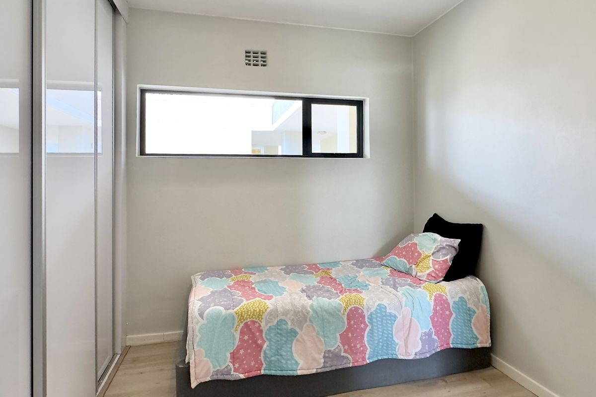 A clean, modern bedroom featuring light-toned flooring, a built-in wardrobe with sliding mirror doors, and a horizontal window for natural light.