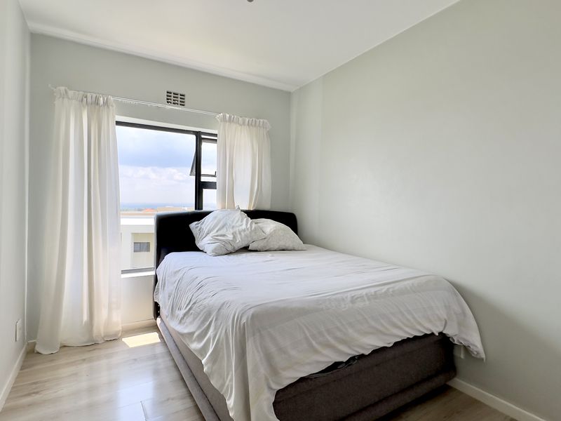 A bright bedroom featuring light-toned laminate flooring, neutral wall finishes, and a window providing natural light and exterior views.