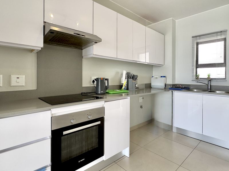 Modern kitchen featuring white gloss cabinetry, stone countertops, integrated electric hob, oven, extractor fan, and a stainless steel sink with tiled flooring.