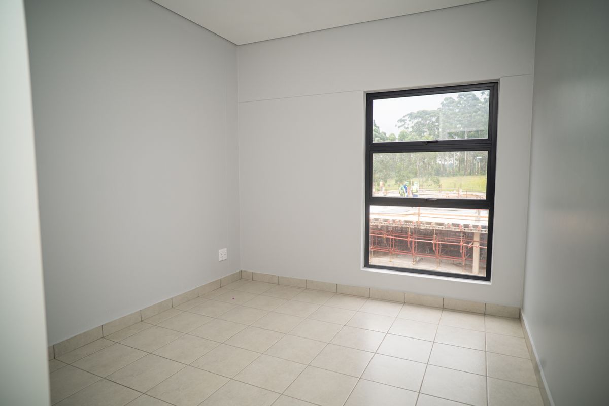 A bright, empty bedroom featuring neutral tiled flooring, smooth grey walls, and a large window with a view of the surrounding area.