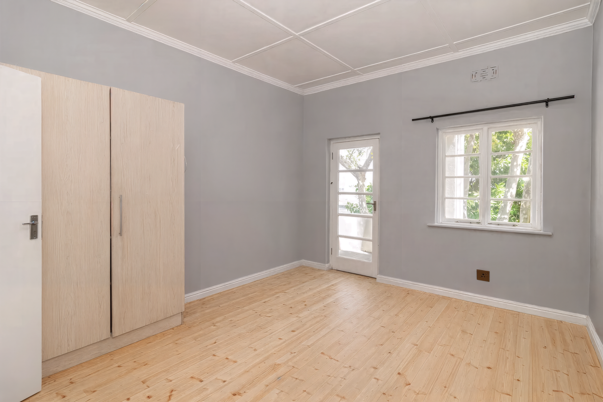 A bright, empty bedroom featuring light wood flooring, built-in cabinetry, a glass-paneled exterior door, and a window with garden views.
