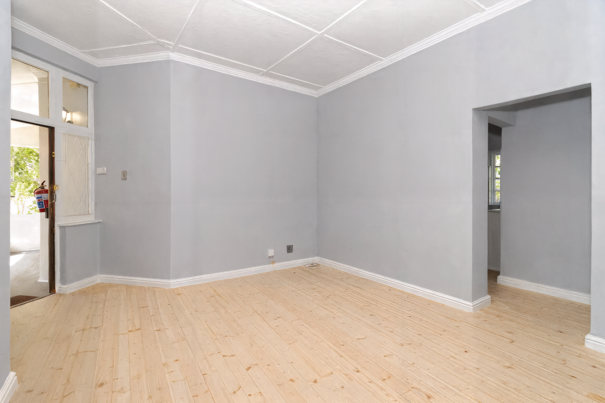 An empty living room featuring light-toned timber flooring, grey walls, and a classic panelled ceiling, with direct access to an outdoor patio area.