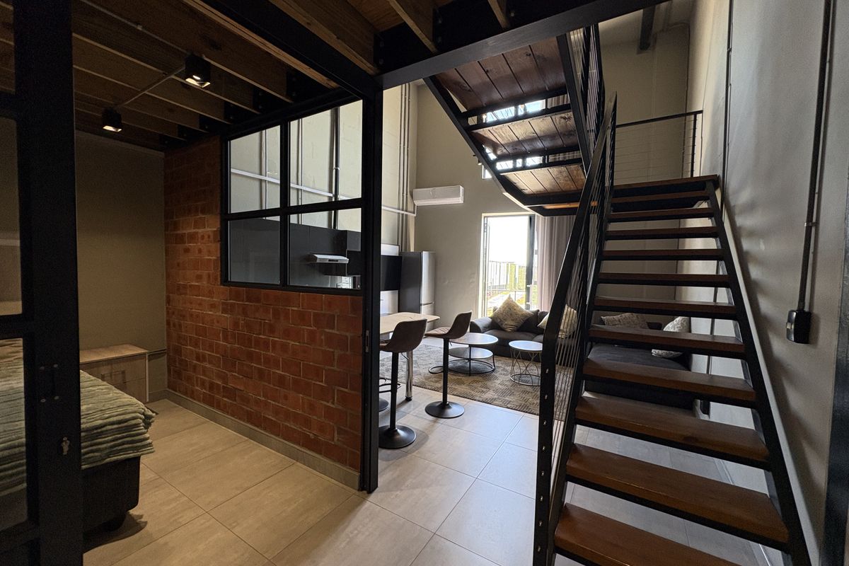 A modern industrial-style apartment featuring an open-plan living area, exposed brick wall, timber staircase, and high ceilings with mezzanine level.