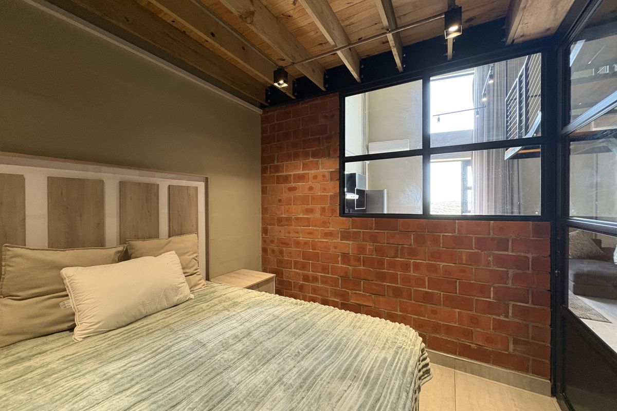 A modern industrial-style bedroom featuring exposed brick walls, timber ceiling beams, and a large black-framed internal window partition.