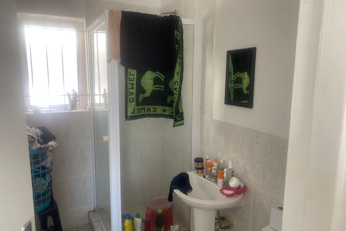 A functional bathroom featuring a corner shower enclosure, pedestal basin, and tiled walls with a security-barred window.