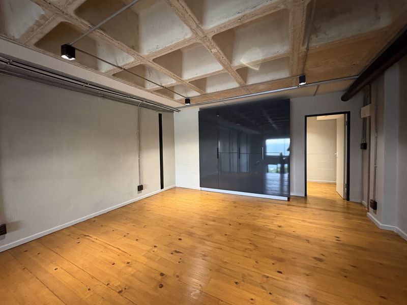A spacious bedroom featuring polished timber flooring, a coffered concrete ceiling with industrial track lighting, and a large built-in wardrobe with a sleek finish.