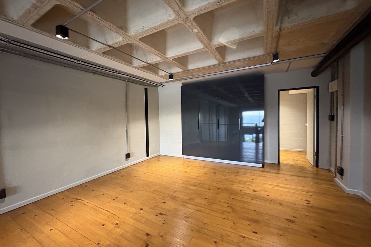 A spacious bedroom featuring polished timber flooring, a coffered concrete ceiling with industrial track lighting, and a large built-in wardrobe with a sleek finish.
